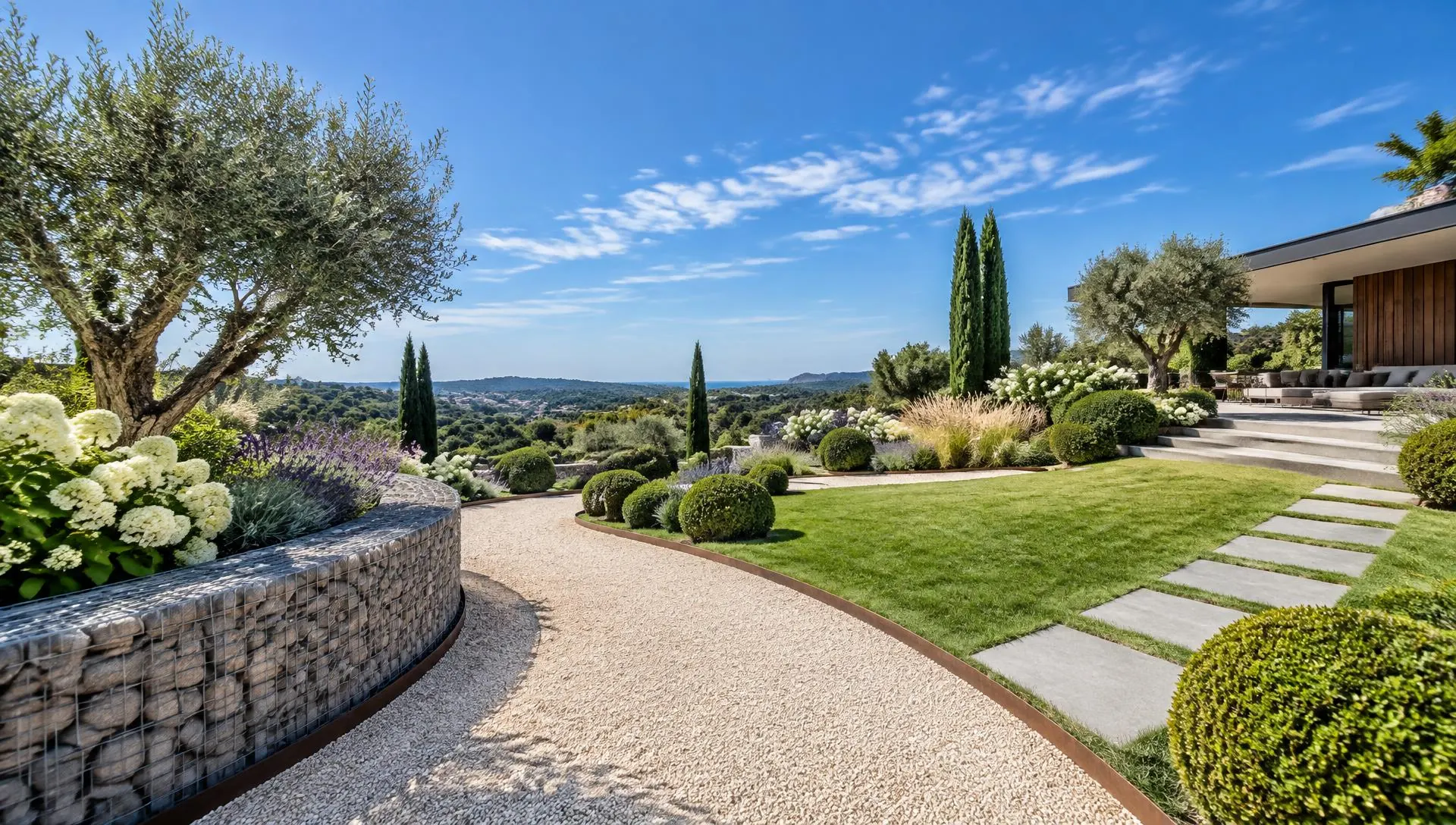 Jardin paysager contemporain : allée de graviers clairs, dalles en pierre grise et mur en gabions sous un ciel bleu